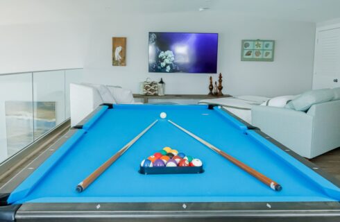 Loft with white walls, hardwood flooring, pool table with blue felt, wall-mounted TV, and upholstered loveseat and chair