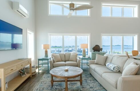 Living room with vaulted ceiling, white walls, hardwood flooring, cream upholstered loveseat and sofa, large wall-mounted TV, and view of marina in the background