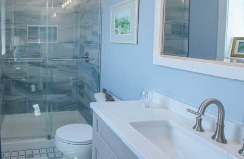 Bathroom with light blue walls, tile flooring, walk-in shower with shades of blue tile, light gray vanity, and white sink