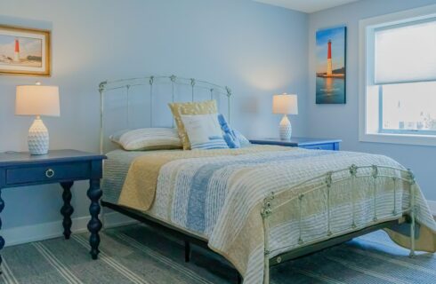 Bedroom with light blue walls, hardwood flooring, white wrought iron bed, yellow, blue, and white comforter, and navy blue tall wooden nightstands with lamps