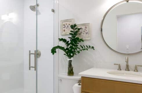 Bathroom with white walls, white-tiled walk-in shower, wooden vanity, white sink, and round mirror