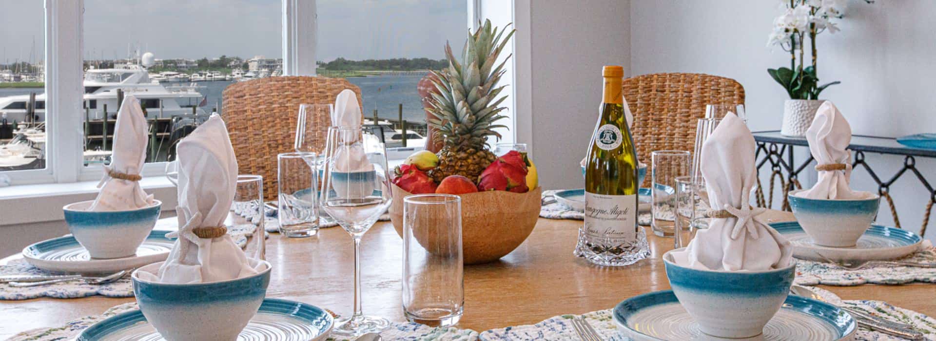 Close up view of wooden dining table with multiple place settings with white and teal plates and bowls, bowl of fruit, and view of marina in the background